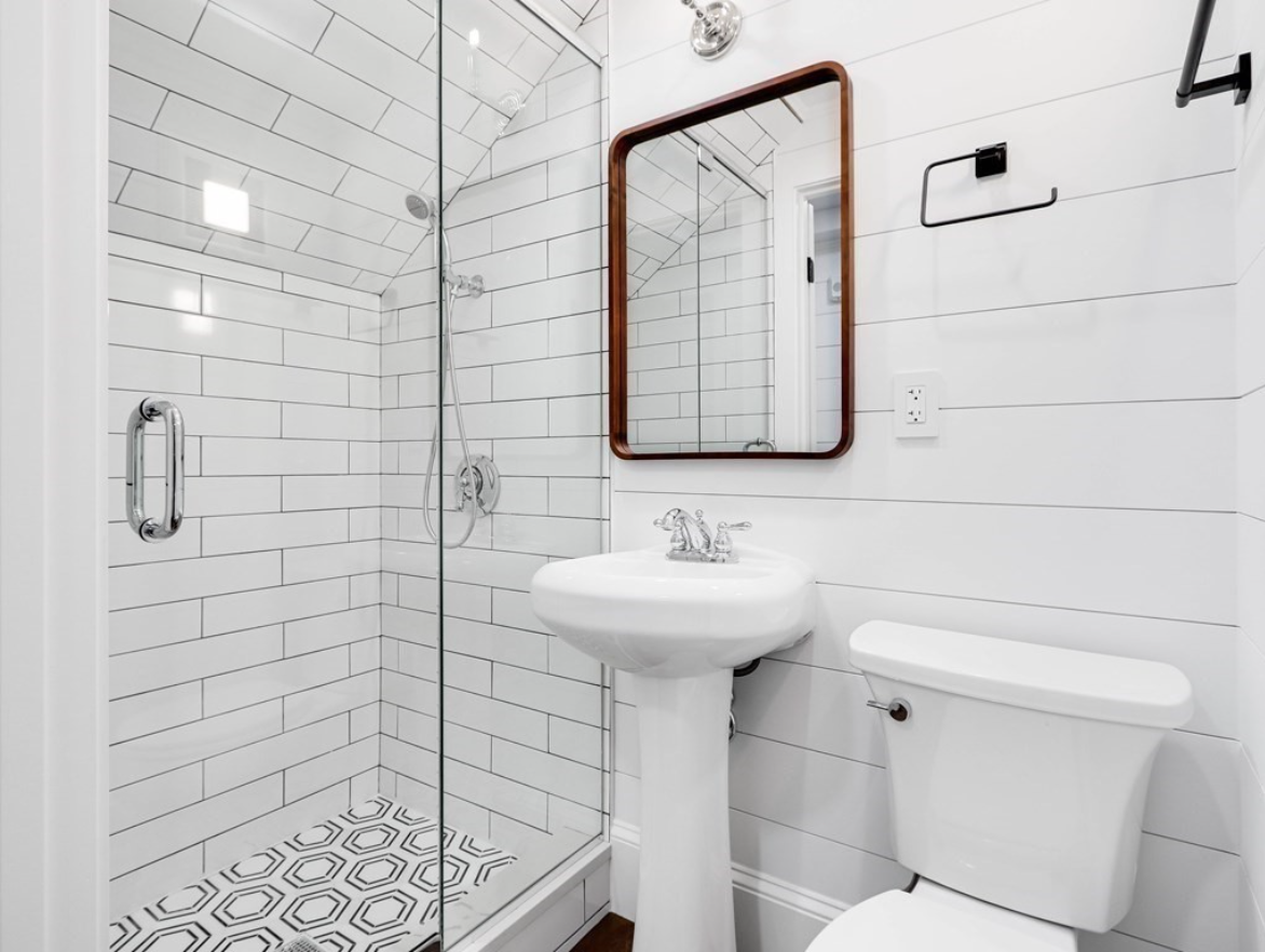 A bathroom with white walls, hardwood floors, and a shower with hexagonal tiling and frameless doors.
