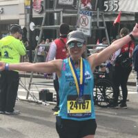 alt = Christine Shearman, posing with her arms up after finishing the 126th Boston Marathon last year. She is wearing a blue Dana-Farber tank, running shorts, a hat, sunglasses, her running bib, and a Boston Marathon medal.
