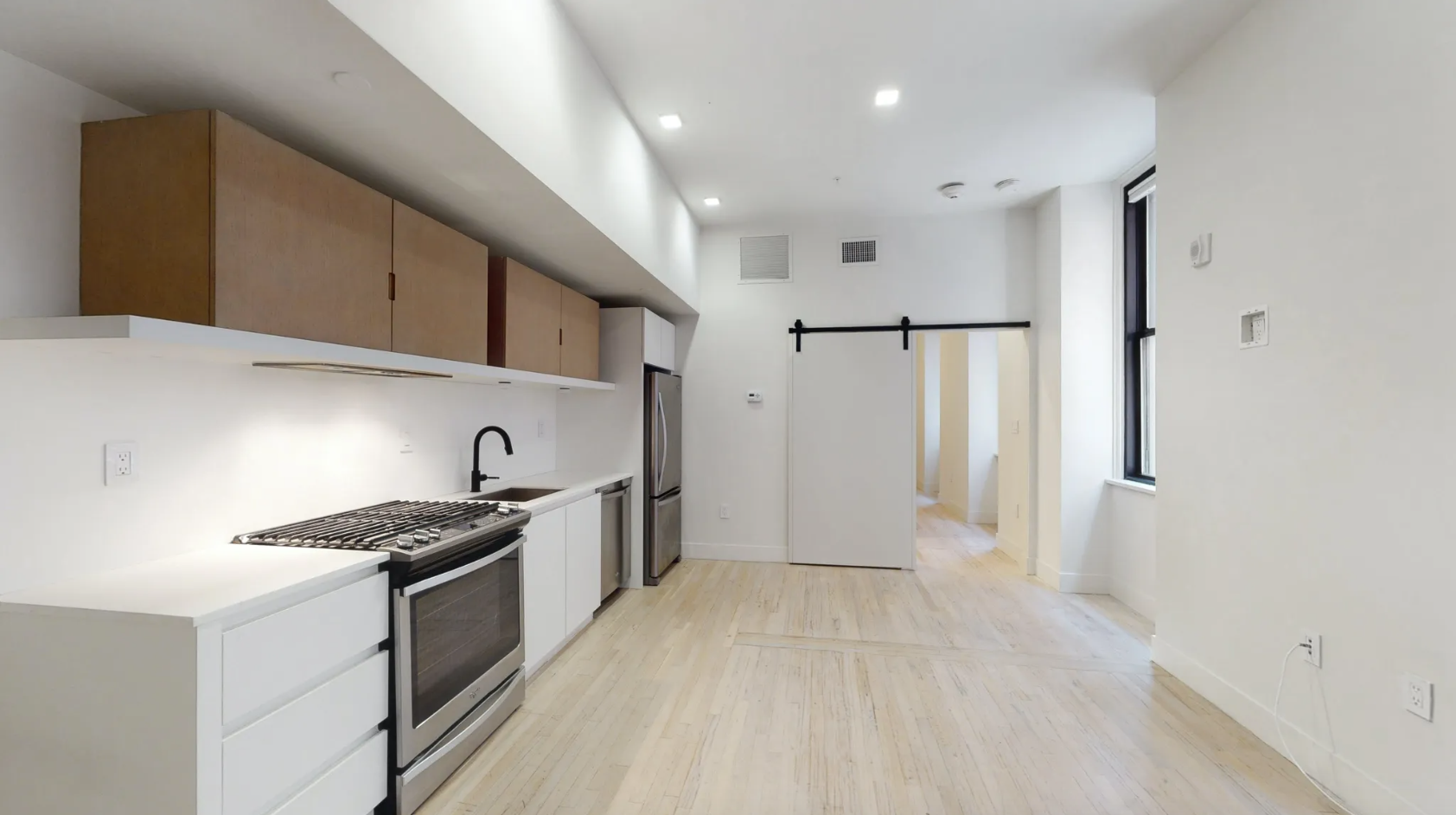 The kitchen has white lower level cabinetry and wooden upper-level cabinetry, hardwood floors, stainless steel appliances, and a barn door into the bedroom.
