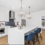 A kitchen with white cabinetry, an island with a white counter that has a waterfall edge. Three stools with cloth seats are parked under two brushed-nickel pendant lights. The stove hood is stainless steel. The backsplash is a navy blue glass tile.