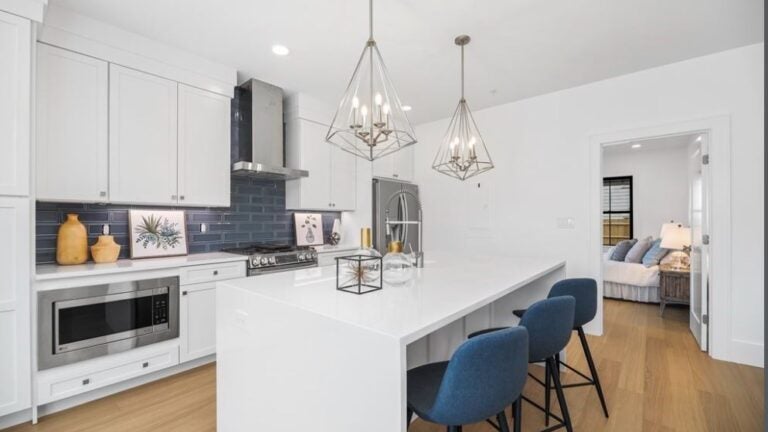 A kitchen with white cabinetry, an island with a white counter that has a waterfall edge. Three stools with cloth seats are parked under two brushed-nickel pendant lights. The stove hood is stainless steel. The backsplash is a navy blue glass tile.