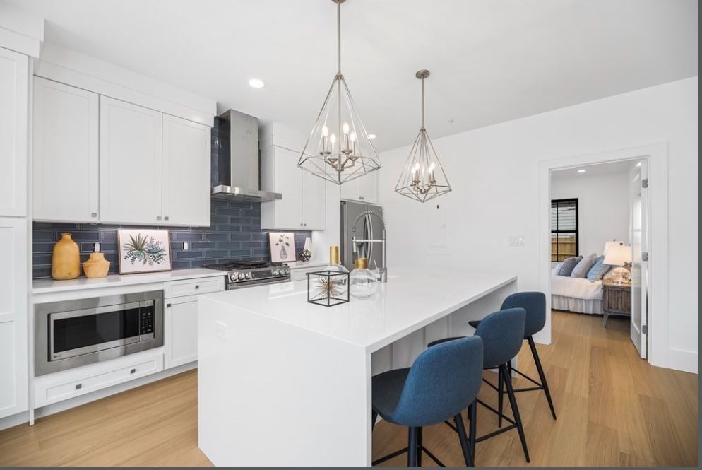 A kitchen with white cabinetry, an island with a white counter that has a waterfall edge. Three stools with cloth seats are parked under two brushed-nickel pendant lights. The stove hood is stainless steel. The backsplash is a navy blue glass tile.