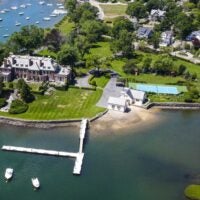 An aerial view of a Georgian estate in Cohasset with an expansive lawn, a long dock, a pool house, tennis courts, a skating pond, and sandy beachfront.