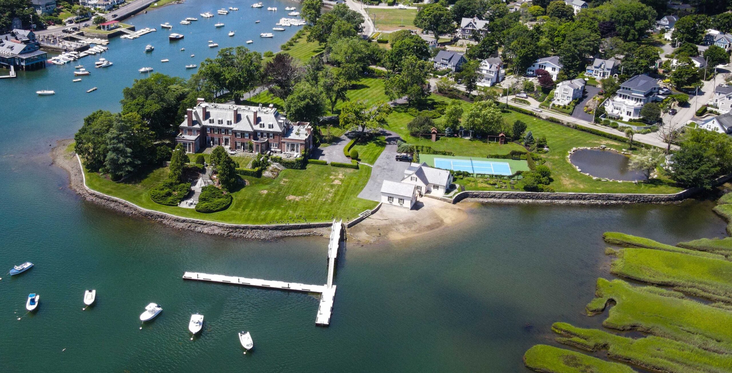 An aerial view of a Georgian estate in Cohasset with an expansive lawn, a long dock, a pool house, tennis courts, a skating pond, and sandy beachfront.