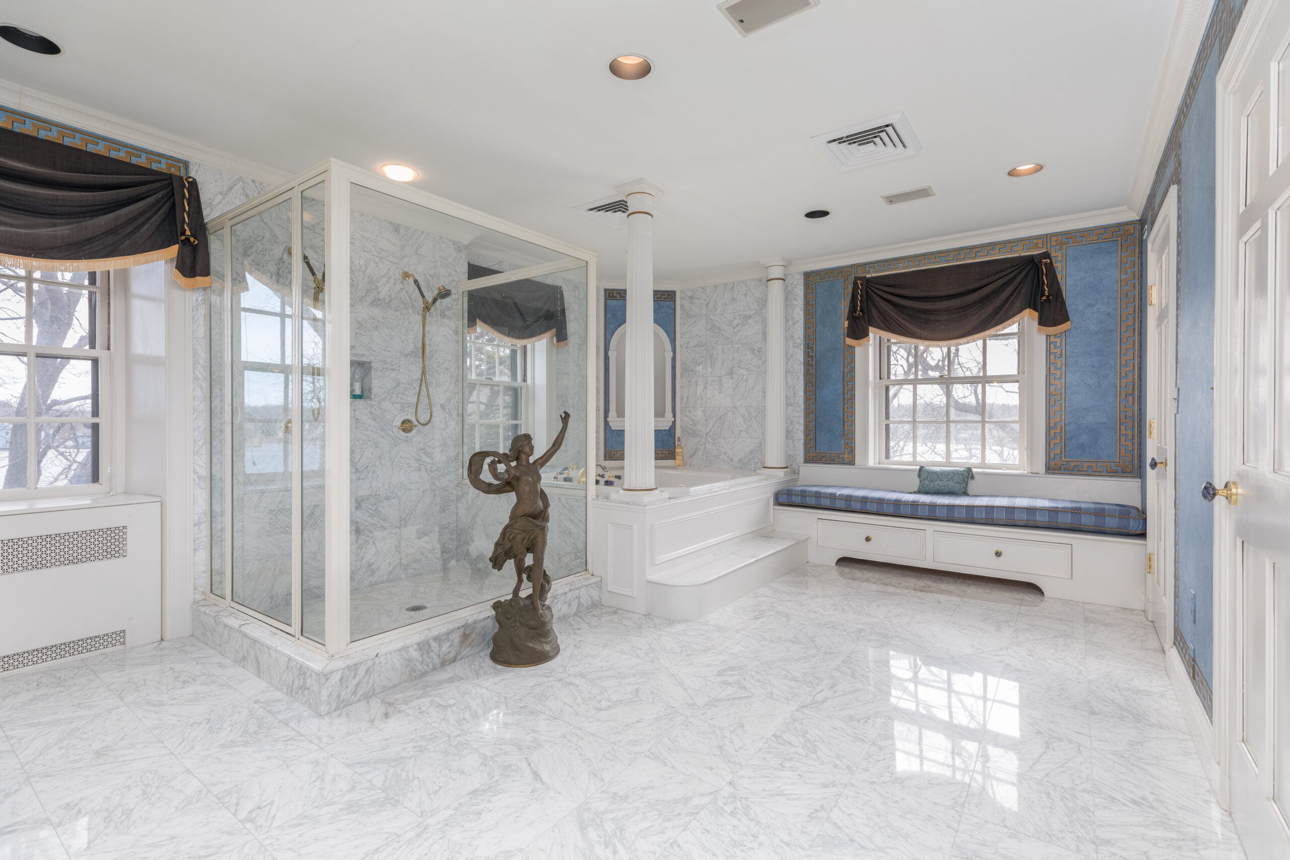 A statue sits in the background of a marble bath with a blue window seat, a walk-in shower, and a soaking tub.