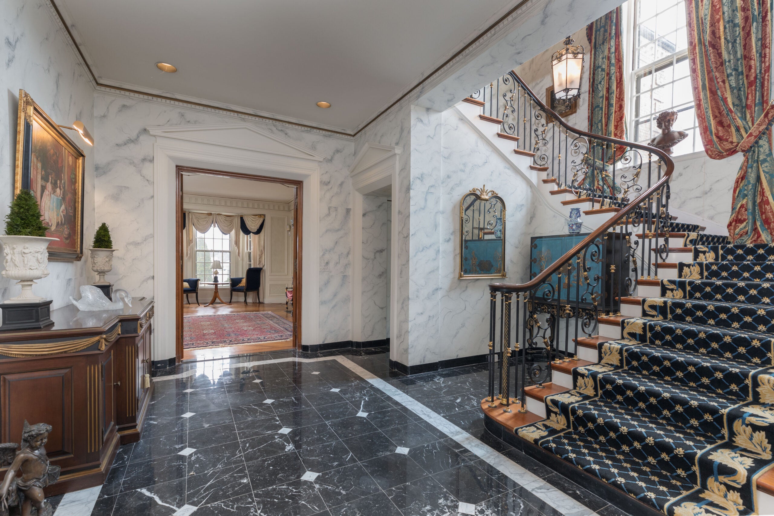 A grand foyer with a black-and-white tile floor, marble walls, and a staircase that cascades up to the right. The trends are covered in a black runner.