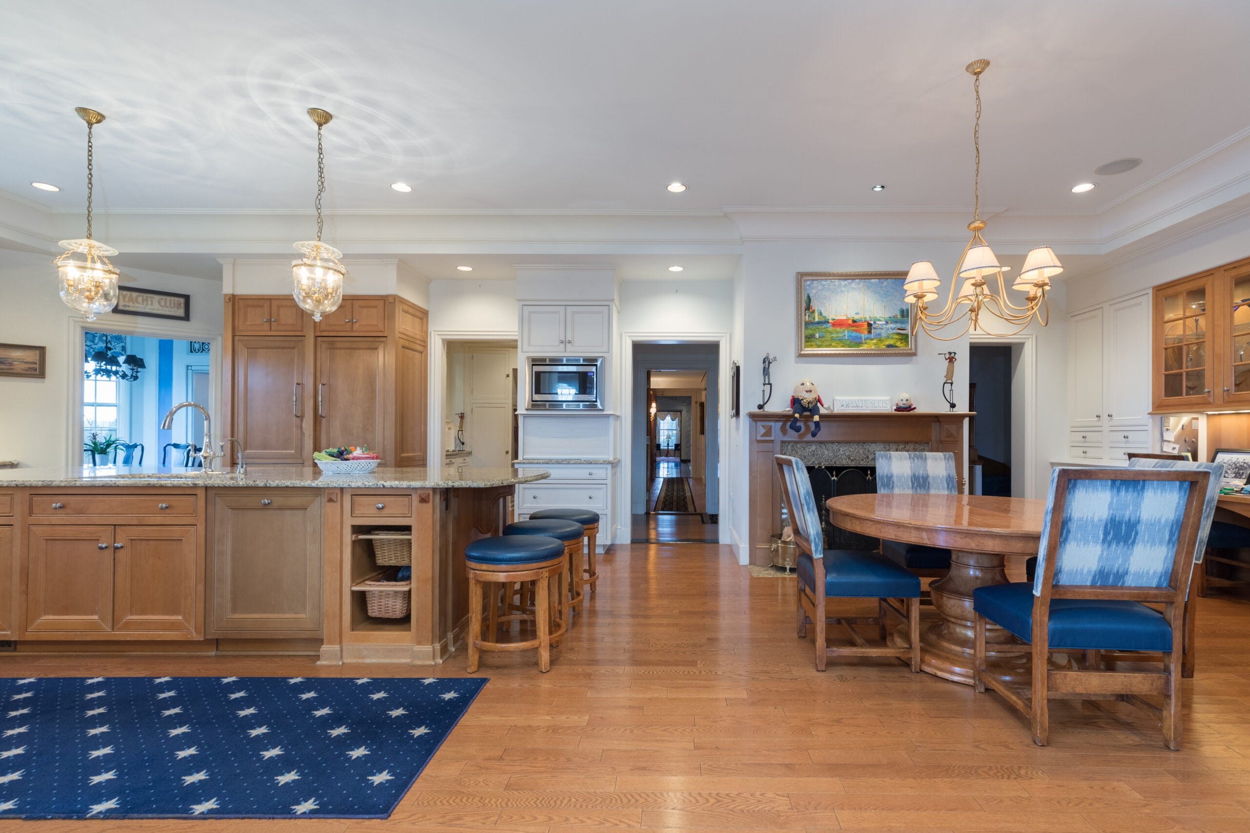 A view of a kitchen with warm-wood cabinetry, a fireplace, a long island with three stools (the cushions are blue), and a round table set before a fireplace.