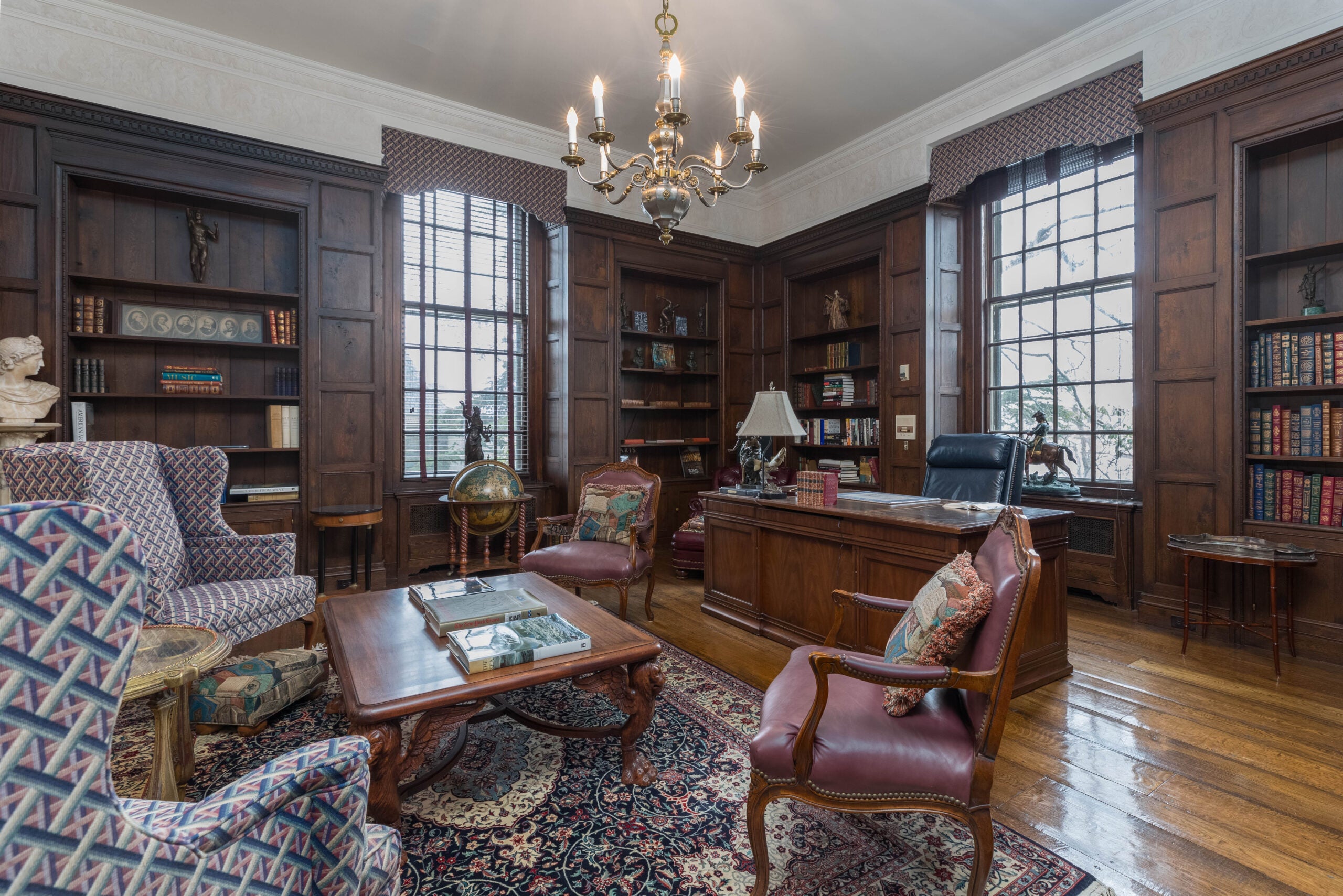 A library with tall windows, wood floors, an Oriental rug, built-in bookshelves, and a candle-like chandelier.