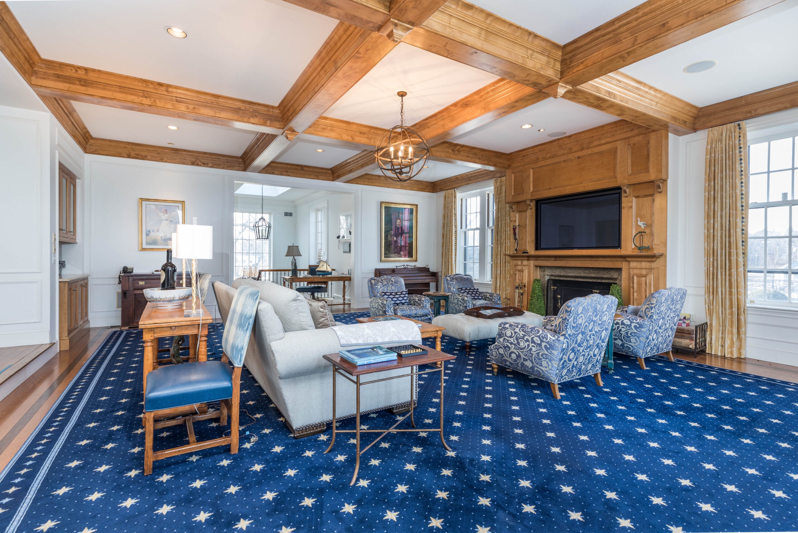 A room filled with overstuffed furniture. The ceiling is coffered with a warm-colored wood, and the floor-to-ceiling fireplace is clad in the same material. The carpeting is white stars on a medium-blue background.