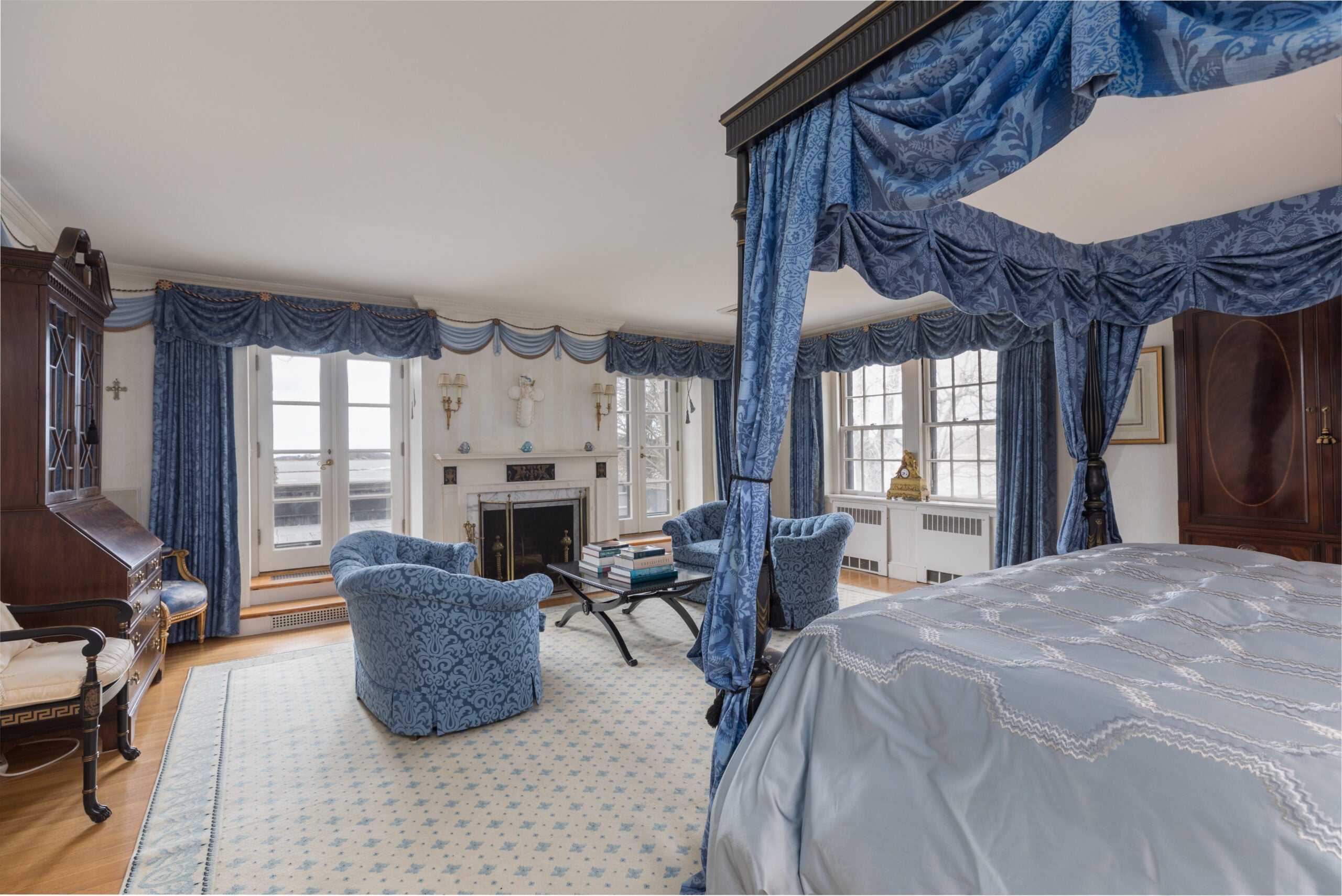 A primary bedroom suite lined with short blue curtains and French doors. A canopy bed sits in the middle with blue drapes. Matching arm chairs are set before the fireplace in this room with light carpeting.