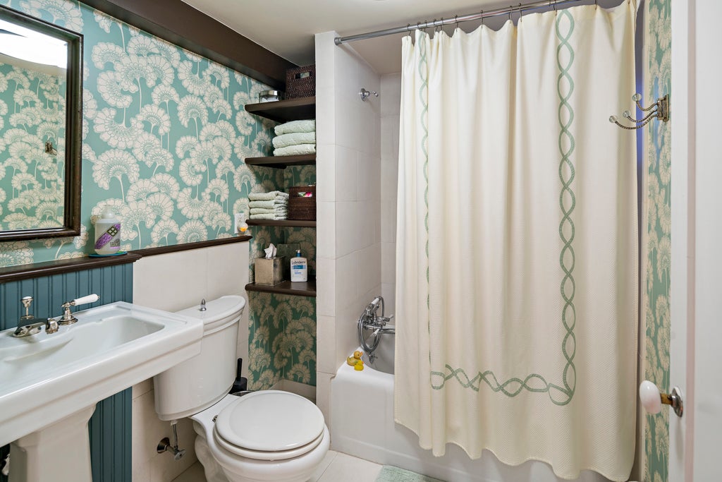 An aqua-colored wallpaper with white dandelion heads covers the walls of this bathroom, which has a white tub/shower combo with a white curtain that has a green chain outline. The toilet and console sink are white.
