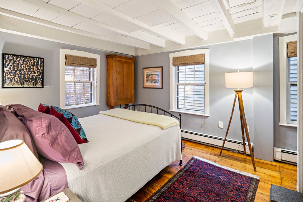 A gray bedroom with a white spindle bed. The bed is clad in white bedding and pink pillows. The room has two windows with wood roll shades. A tripod lamp sits in the corner. The room has exposed white beams.