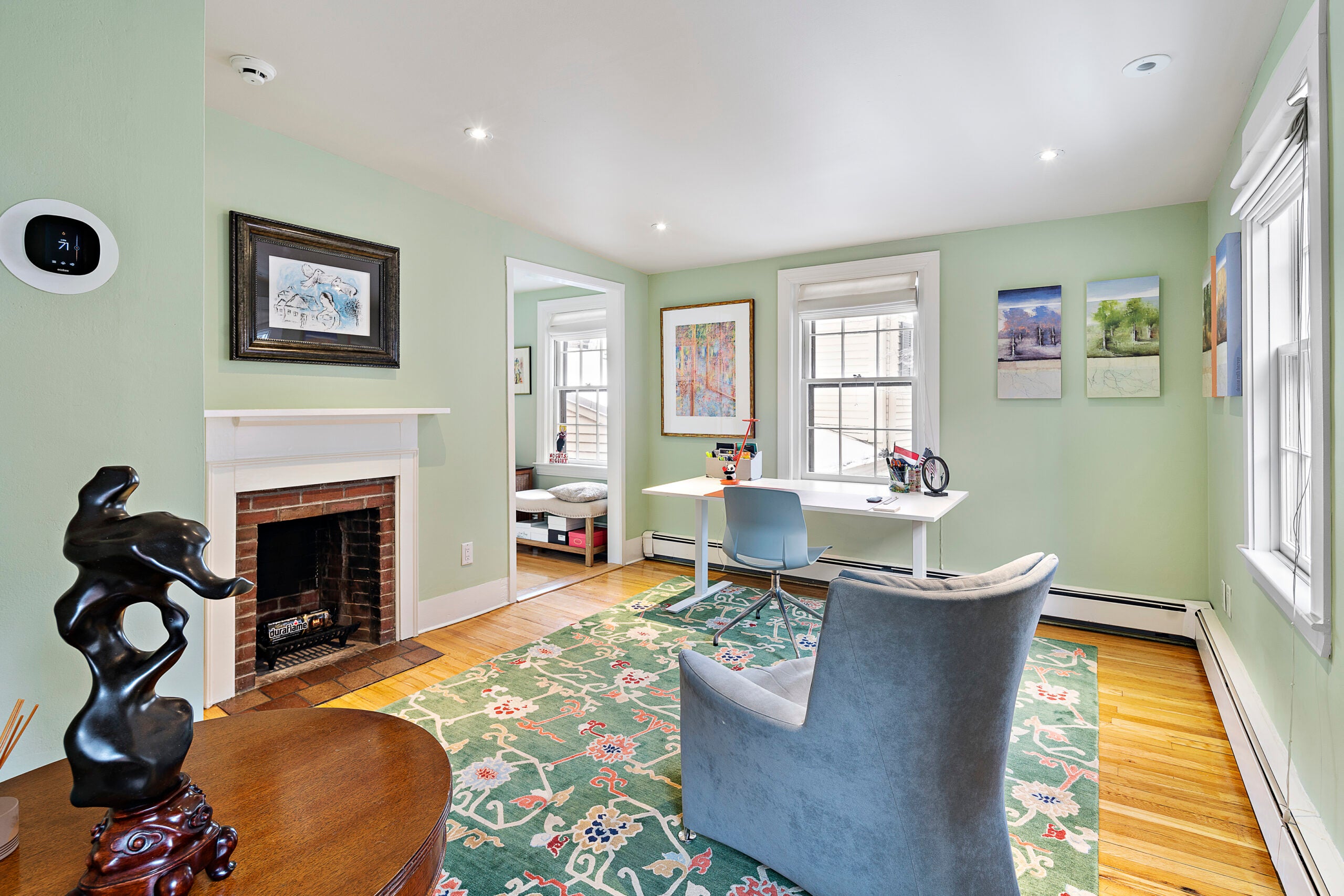 A green living room with a white fireplace, recessed lighting, hardwood flooring,a green floral rug, a window, a narrow desk before the window with a blue chair, and a blue arm chair.