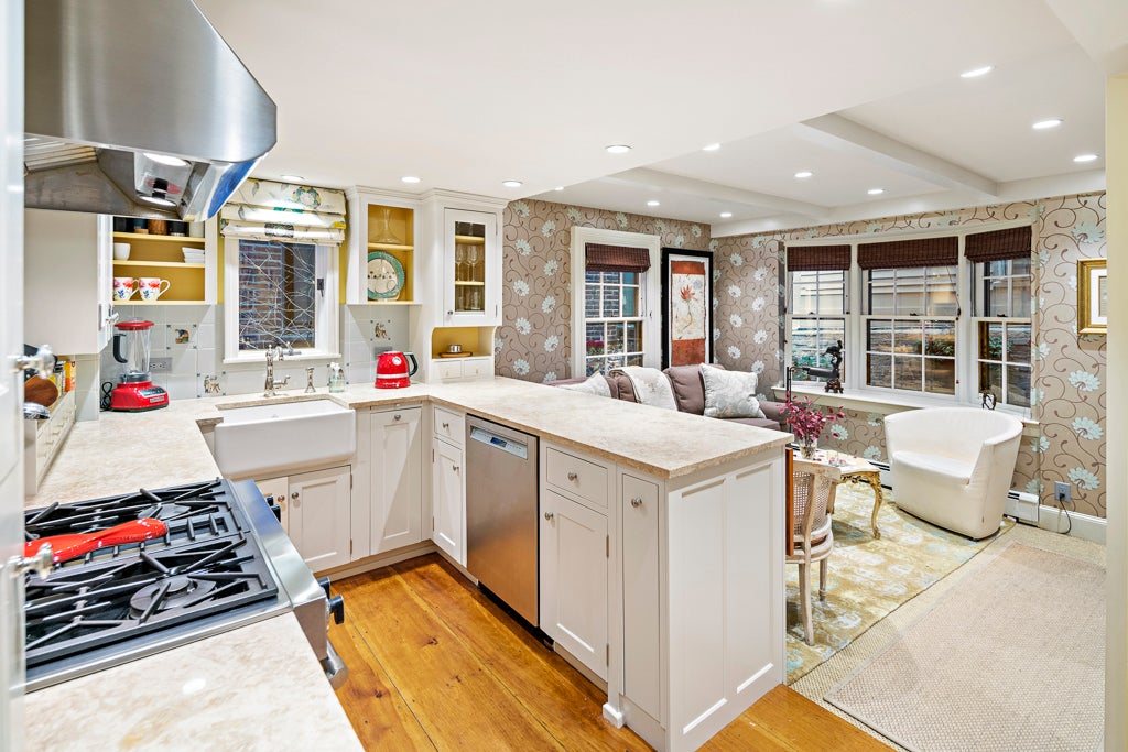 A kitchen with white cabinets, a farmhouse sink, and a peninsula. The kitchen overlooks a living room area with tan wallpaper with a floral pattern.