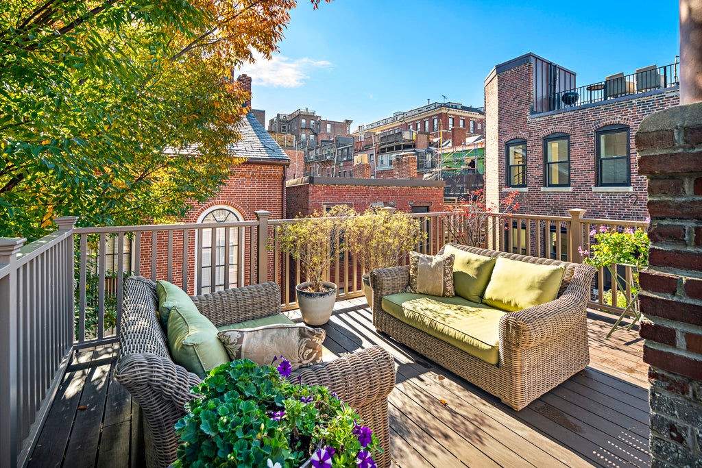 A roof deck overlooking brick buildings. The sky is blue and fairly cloudless. A basket of purple petunias cascades in the foreground. The deck has wicker furniture with light green cushions.