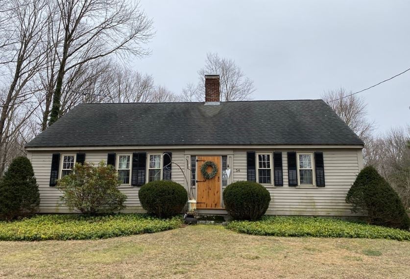 The exterior of a gray antique Cape with five windows, a wooden door, and central chimney, black shutters, a black roof, and two patches of ground cover next to four shrubs.