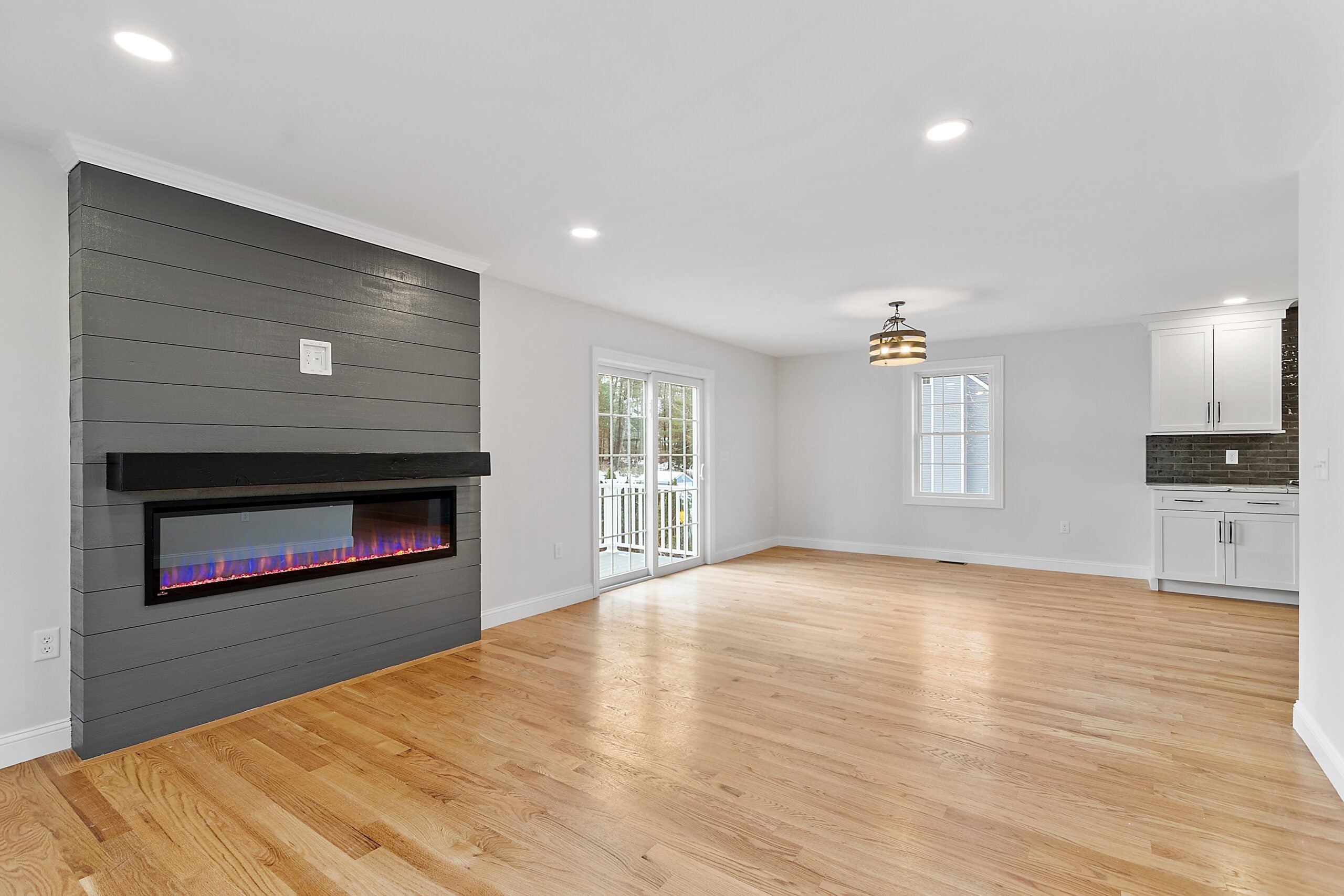 A view of a fireplace that extends to the ceiling. It is electric with a long, narrow glass screen. The fireplace is clad in a charcoal-colored material. The room has recessed lighting and hardwood flooring, and one can see a drum-shade light fixture, a slider, kitchen cabinetry, and a window in the background.