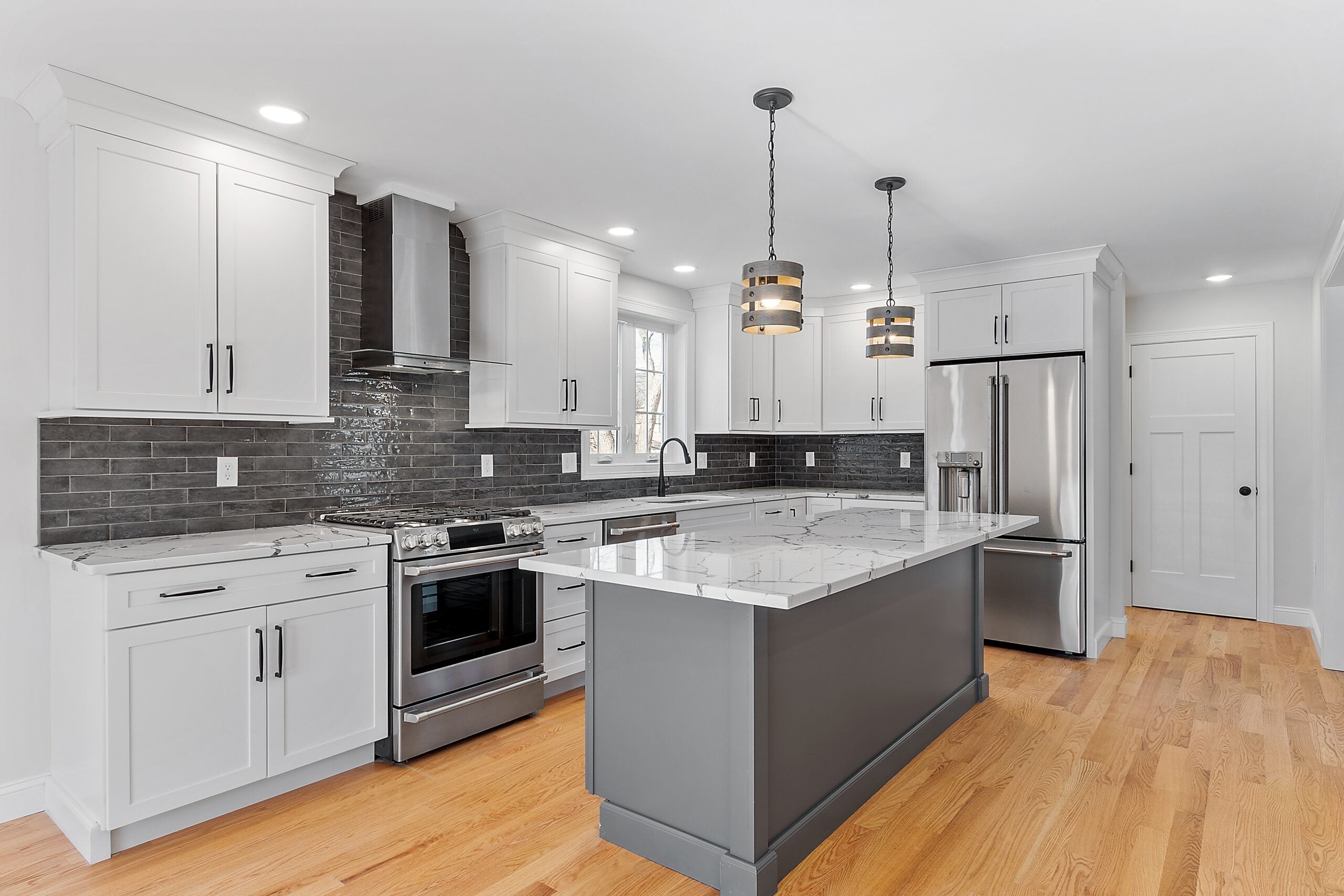 A longways view of a kitchen with a gray island with a light-colored countertop, white cabinets with black handles, a dark glass tile backsplash, and two pendant lights with shades that look like straps of wood.