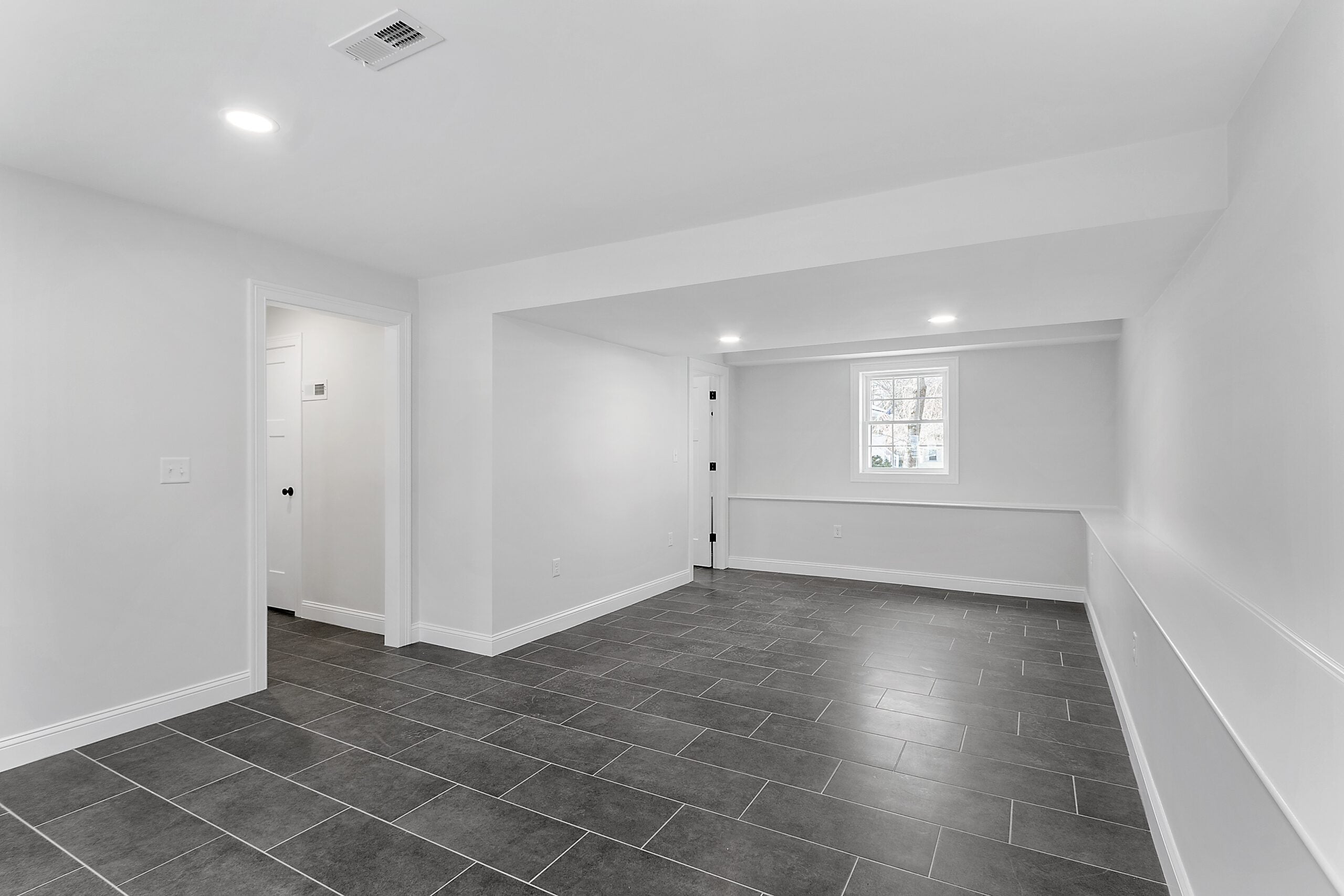 A room with a small window, chair rail molding, wide gray porcelain tile flooring, recessed lighting, and white walls.