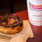 A white to-go coffee cup with a Dunkin' logo sits on a table next to a chocolate-frosted doughnut covered in rainbow sprinkles.
