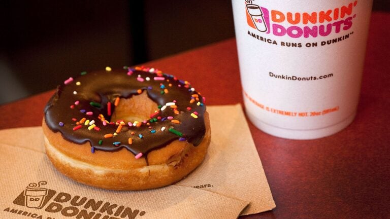A white to-go coffee cup with a Dunkin' logo sits on a table next to a chocolate-frosted doughnut covered in rainbow sprinkles.