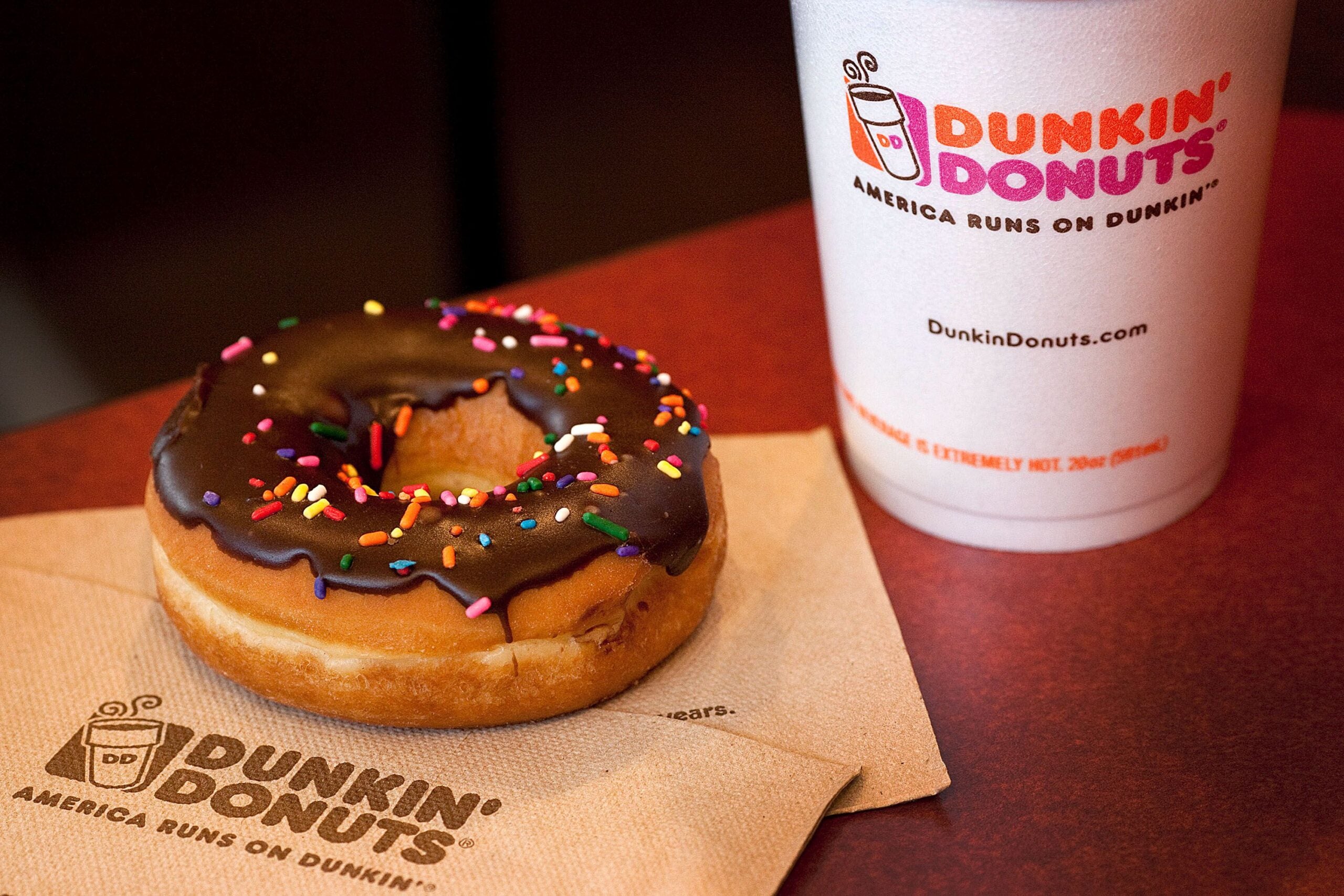 A white to-go coffee cup with a Dunkin' logo sits on a table next to a chocolate-frosted doughnut covered in rainbow sprinkles.