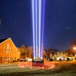 Skylights on the lawn outside Holy Family Church in Duxbury project into the nighttime sky. Three of the four beams are white light, and one is purple.