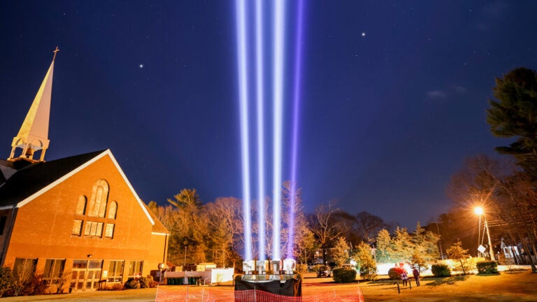 Skylights on the lawn outside Holy Family Church in Duxbury project into the nighttime sky. Three of the four beams are white light, and one is purple.