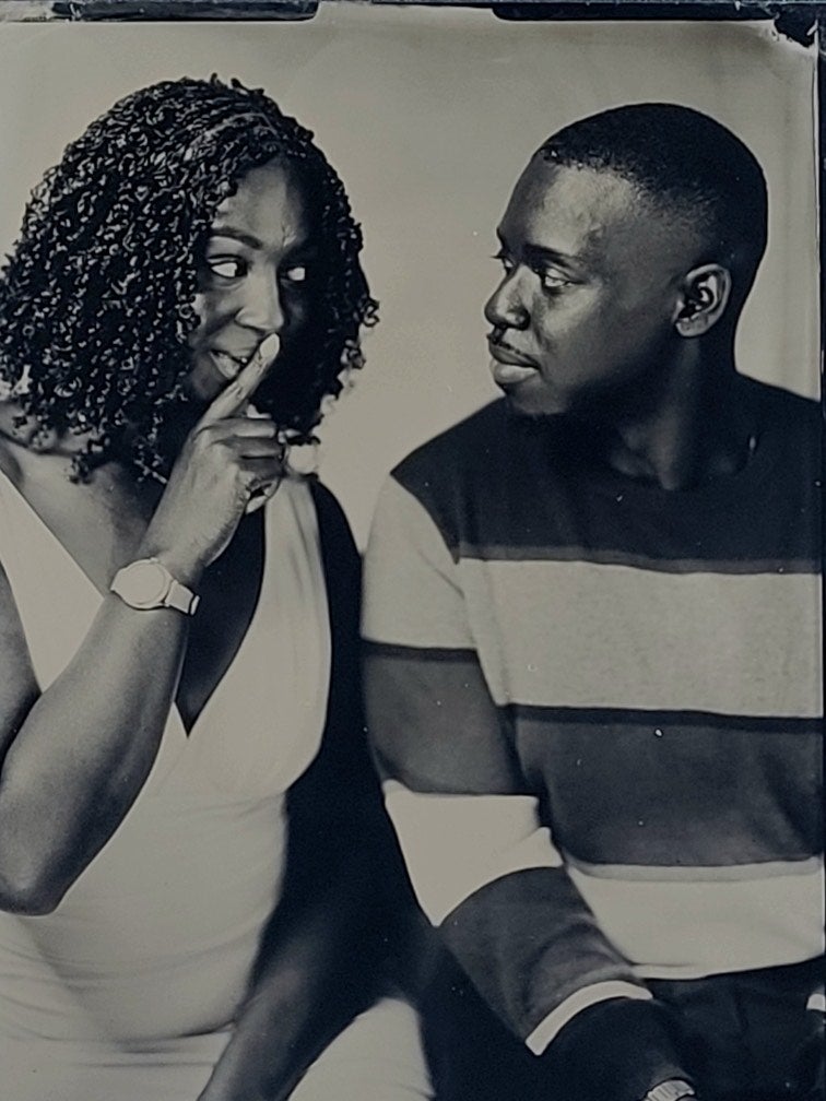 A black-and-white photograph of a woman and a man. The woman is holding a finger to her lips while the man turns to look her in the eyes.