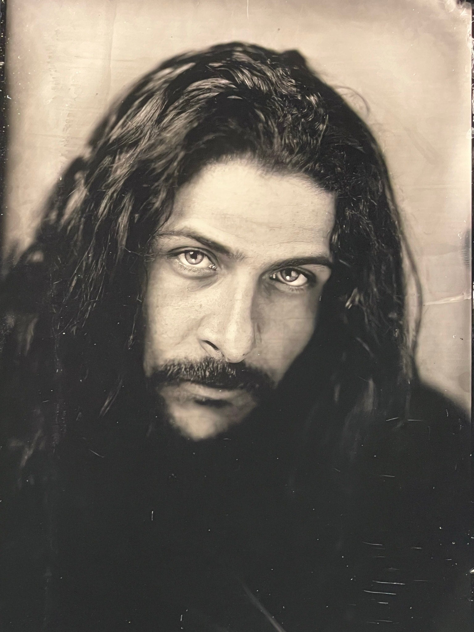 A black-and-white tintype photo of a man with long hair and a facial hair, staring solemnly into the camera.