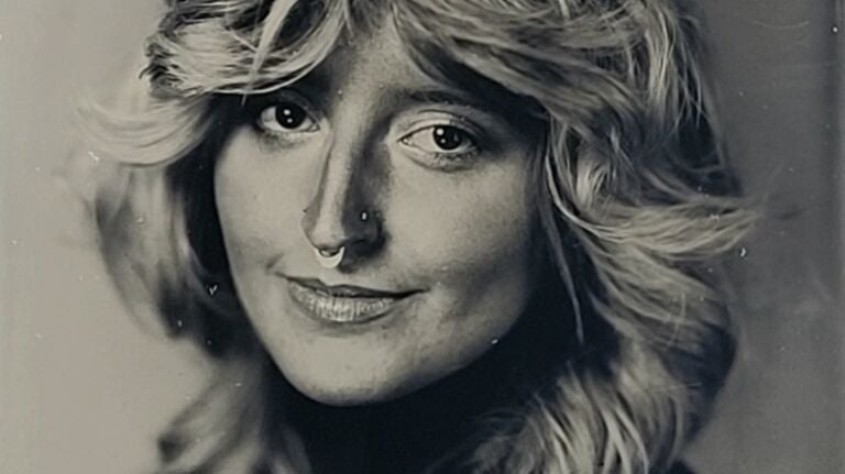 A woman is shown in a black-and-white tintype with long, feathery hair, a small smile, and nose piercings.