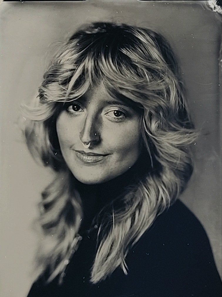 A woman is shown in a black-and-white tintype with long, feathery hair, a small smile, and nose piercings.