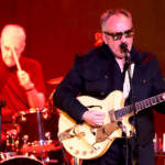 Elvis Costello dressed in black playing his guitar, with drummer Pete Thomas in the background