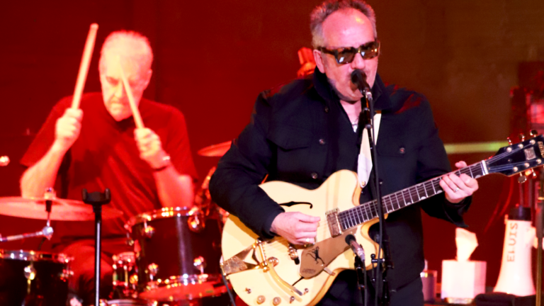Elvis Costello dressed in black playing his guitar, with drummer Pete Thomas in the background