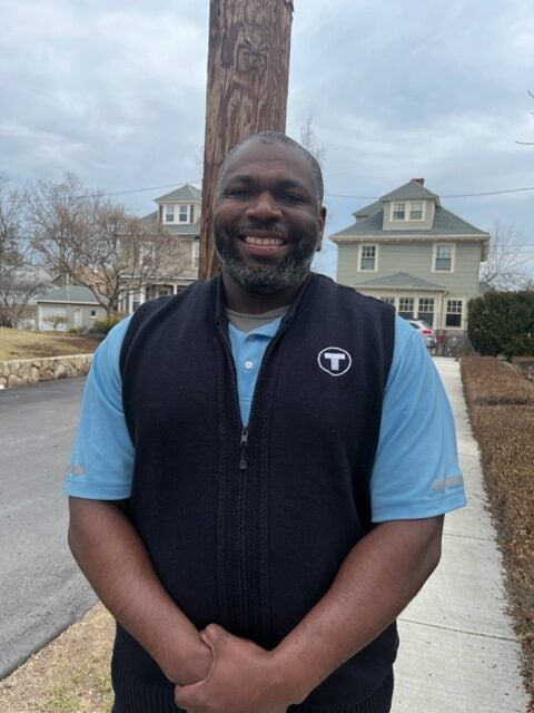 Green Line T driver Wali Holly, a middle-aged Black man shown smiling outdoors in a residential area, wearing in a blue short-sleeved shirt and a black vest with the white T logo on the chest.