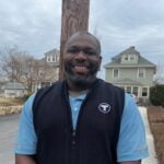 Green Line T driver Wali Holly, a middle-aged Black man shown smiling outdoors in a residential area, wearing in a blue short-sleeved shirt and a black vest with the white T logo on the chest.