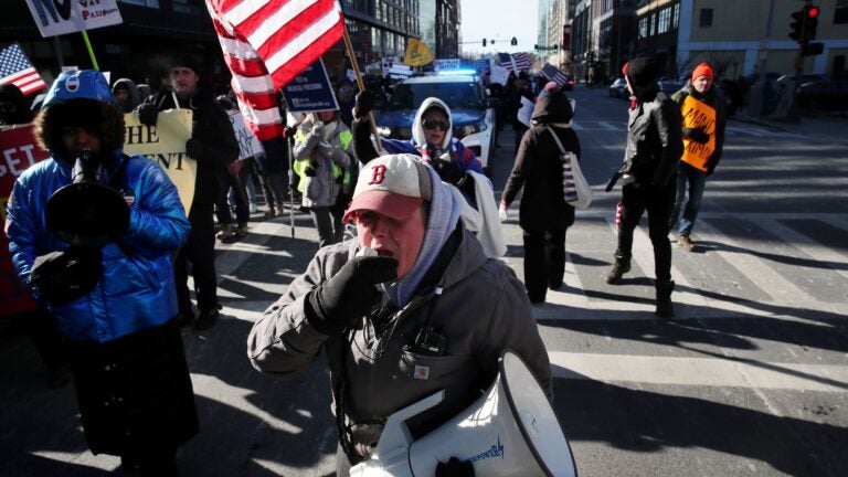 Former Boston police Sergeant Shana Cottone is shown holding a megaphone and protesting in the street, wearing winter clothing, with an American flag behind her.