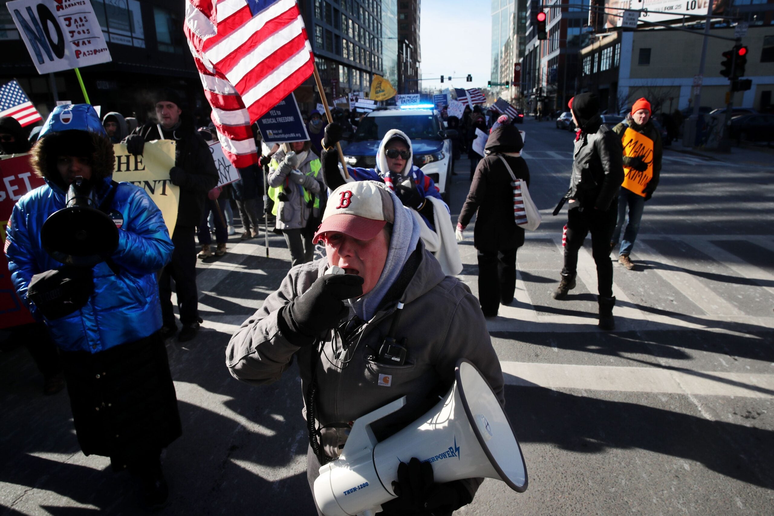 Former Boston police Sergeant Shana Cottone is shown holding a megaphone and protesting in the street, wearing winter clothing, with an American flag behind her.