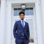 A man wearing a blue suit stands in front of a white door.