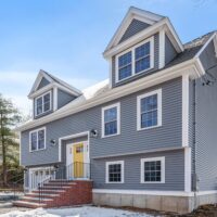 A angled view of the Home of the Week: A gray, dormered Cape with a bright yellow front door and brick front steps.
