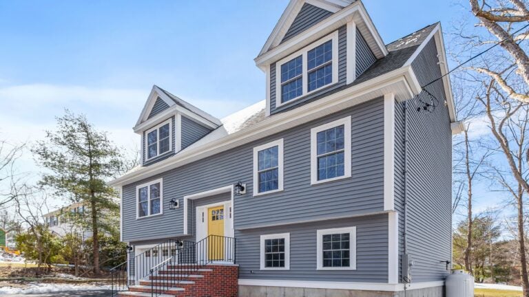 A angled view of the Home of the Week: A gray, dormered Cape with a bright yellow front door and brick front steps.