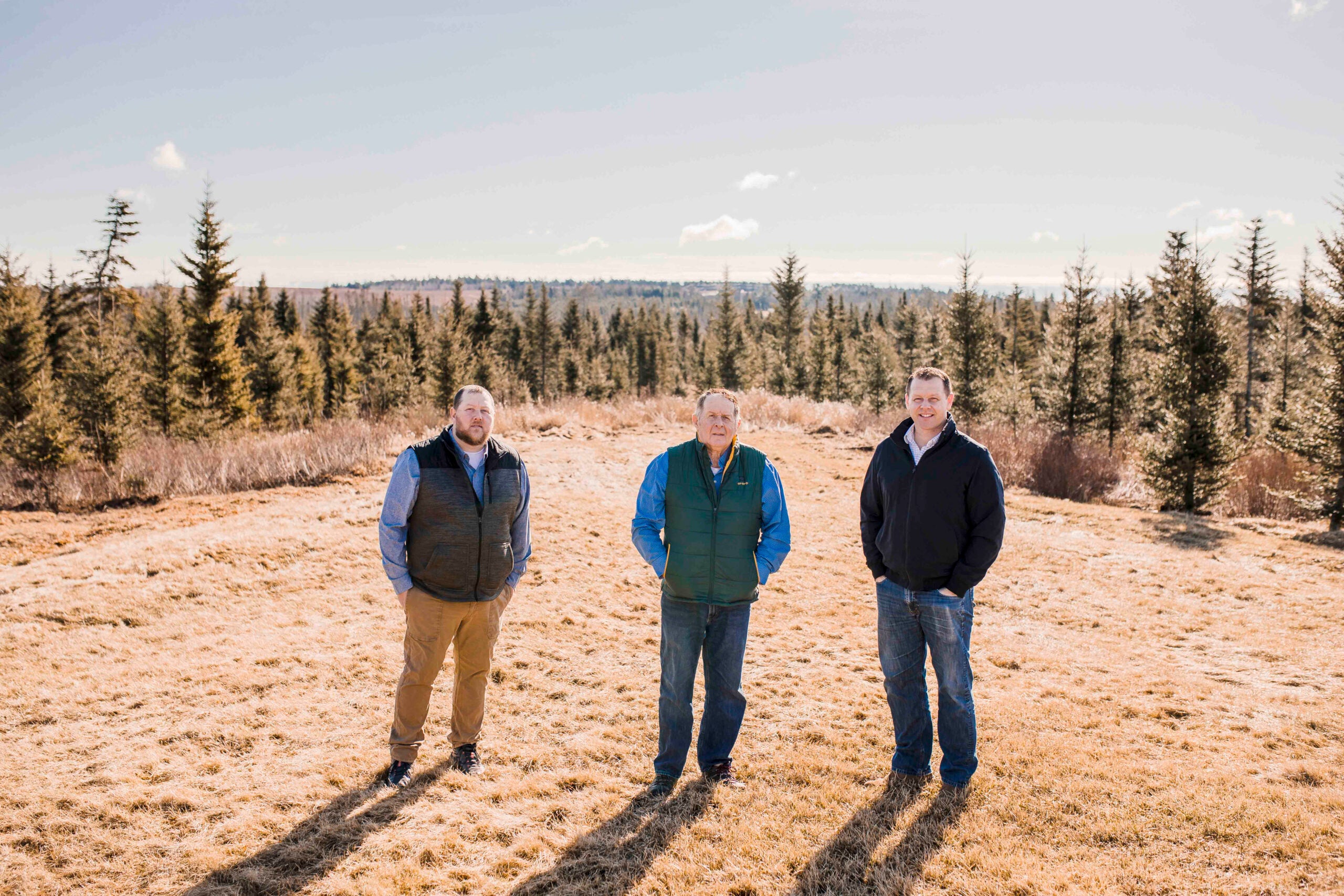 Mike, Morrill, and Rob Worcester stand outside in an open, grassy area with a wooded expanse behind them.