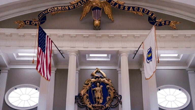 The seal of the Commonwealth of Massachusetts, featuring a Native American at center, is displayed on a statue and flag