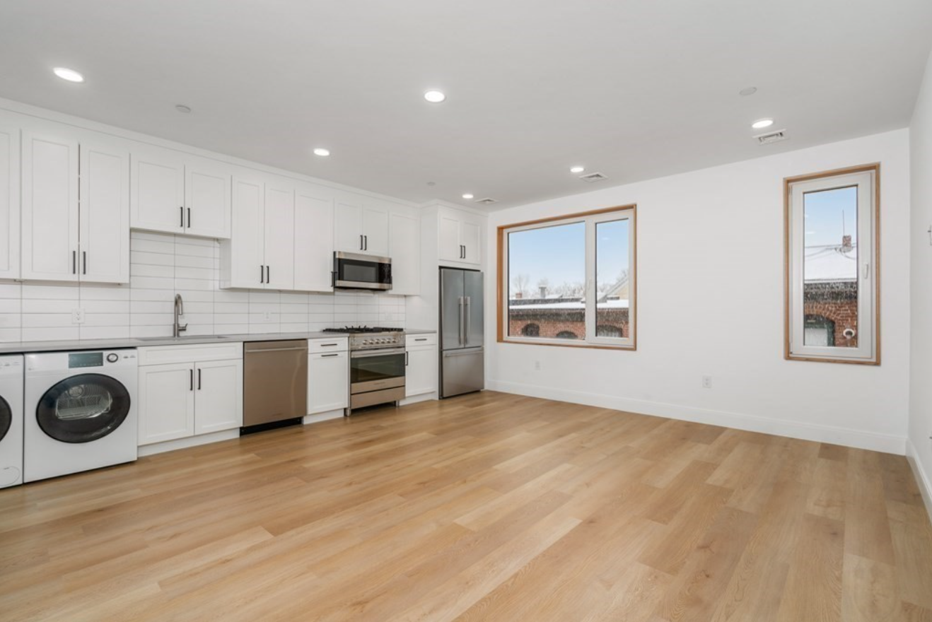 The studio condo for sale has hardwood floors, white walls, and wood-framed windows. The kitchen area features stainless steel appliances, white Shaker-style cabinets, and a washer and dryer.