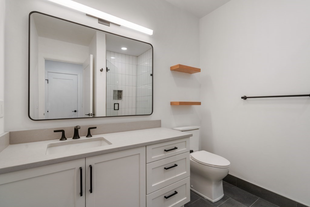 The bathroom in this condo for sale has a single vanity, floating wood shelves, and a walk-in shower with vertically-stacked subway tile.