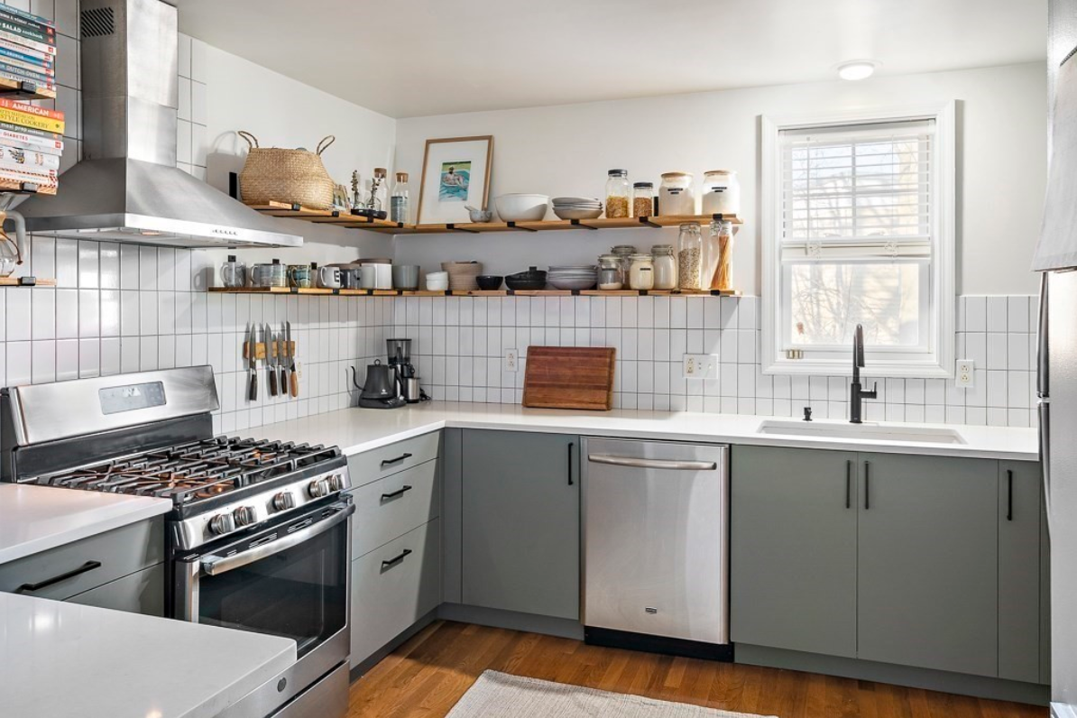 Floating shelves provide storage space, and a single-hung window lets light into the kitchen. Vertically-stacked subway tile backsplash is set above gray flat-panel cabinets and stainless steel appliances.