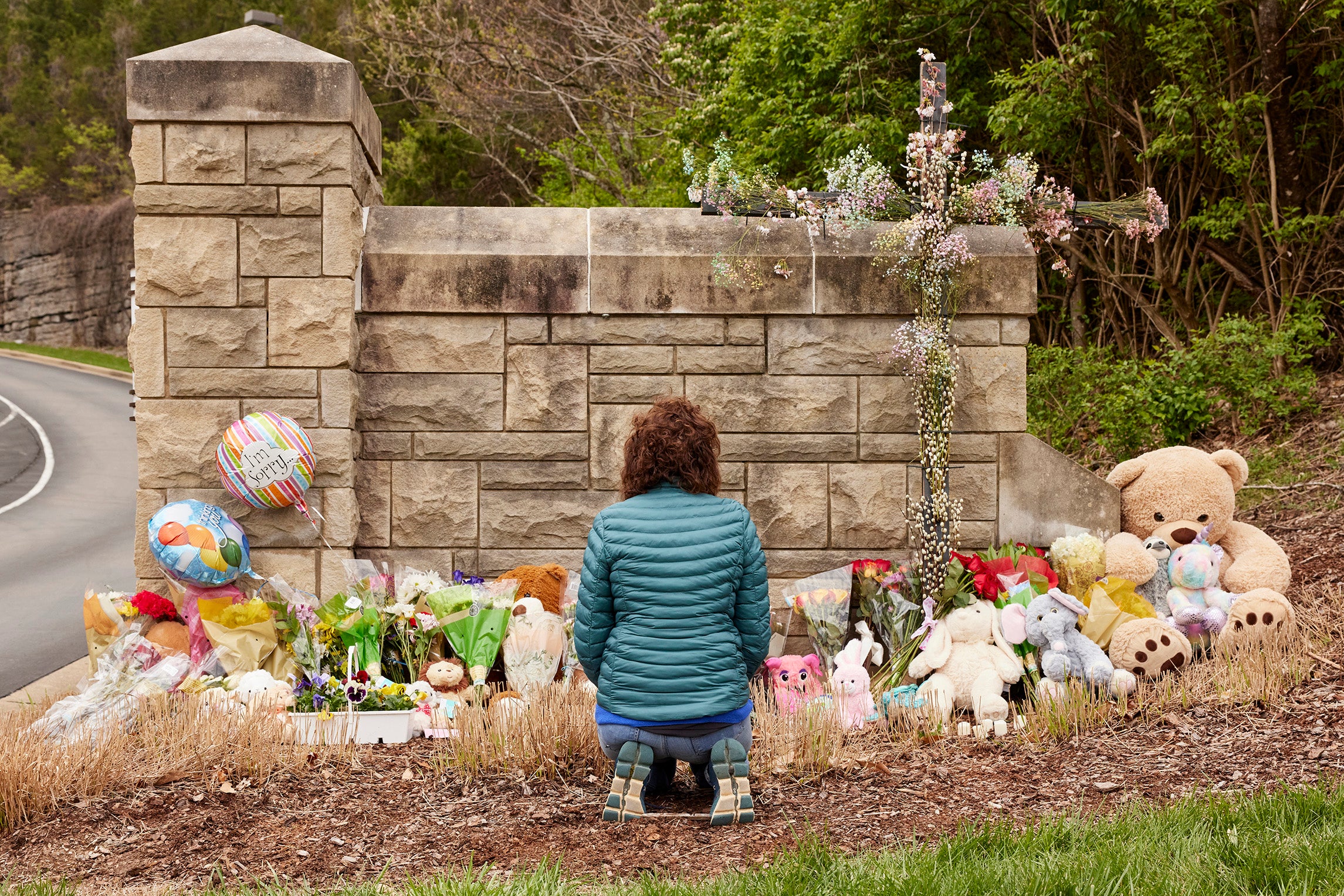 A woman in a teal jacket with brown hair kneels at a makeshift memorial of flowers and toys placed in front of a stone wall.