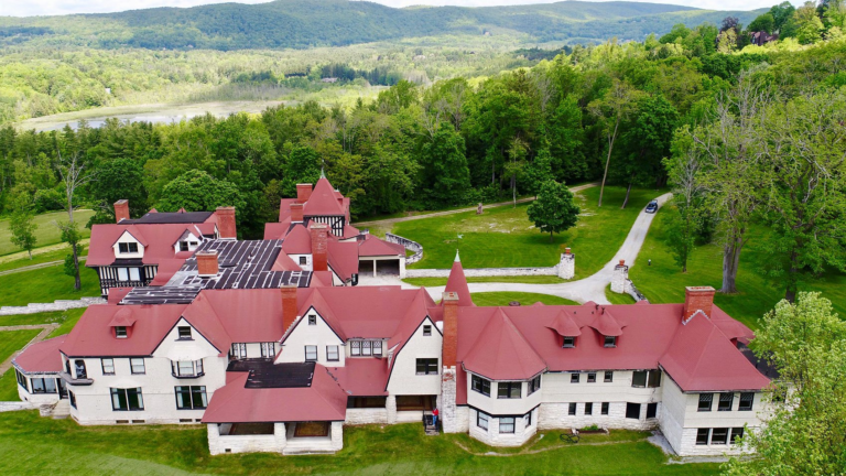A view of a red-roofed Berkshires mansion with a myriad of rooflines and turrets. The walls are white, and the Berkshires hills are visible in the background.