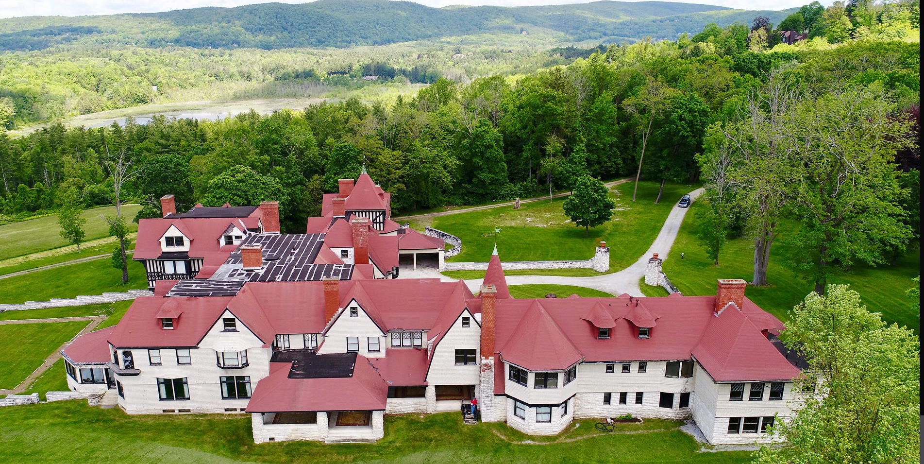 A view of a red-roofed Berkshires mansion with a myriad of rooflines and turrets. The walls are white, and the Berkshires hills are visible in the background.
