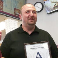 Stash's Pizza owner Stavros Papantoniadis is shown wearing a black collared shirt and holding up an "A" grade sanitary inspection certificate.